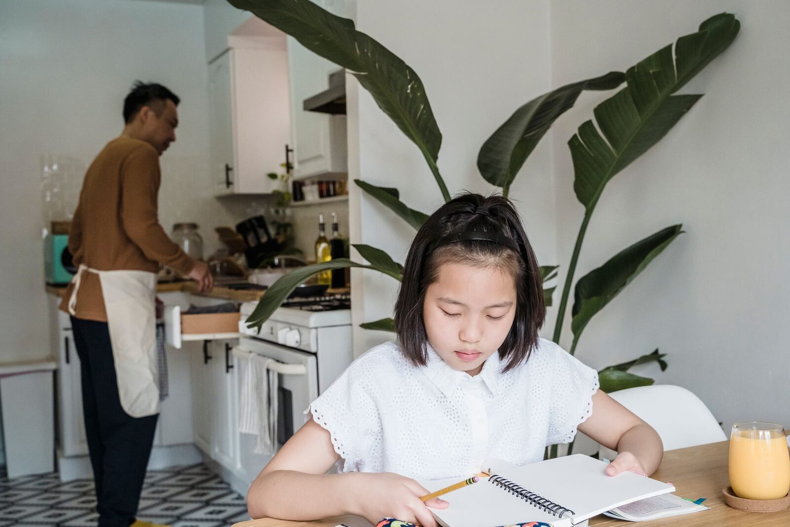 A girl does her homework at the kitchen table while a parent cooks nearby